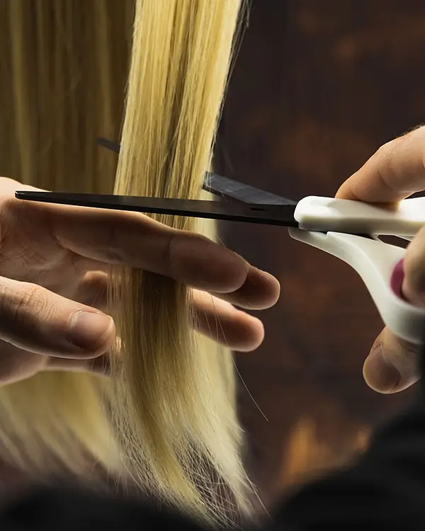 Hands holding a section of straight blonde hair while scissors trim the ends.
