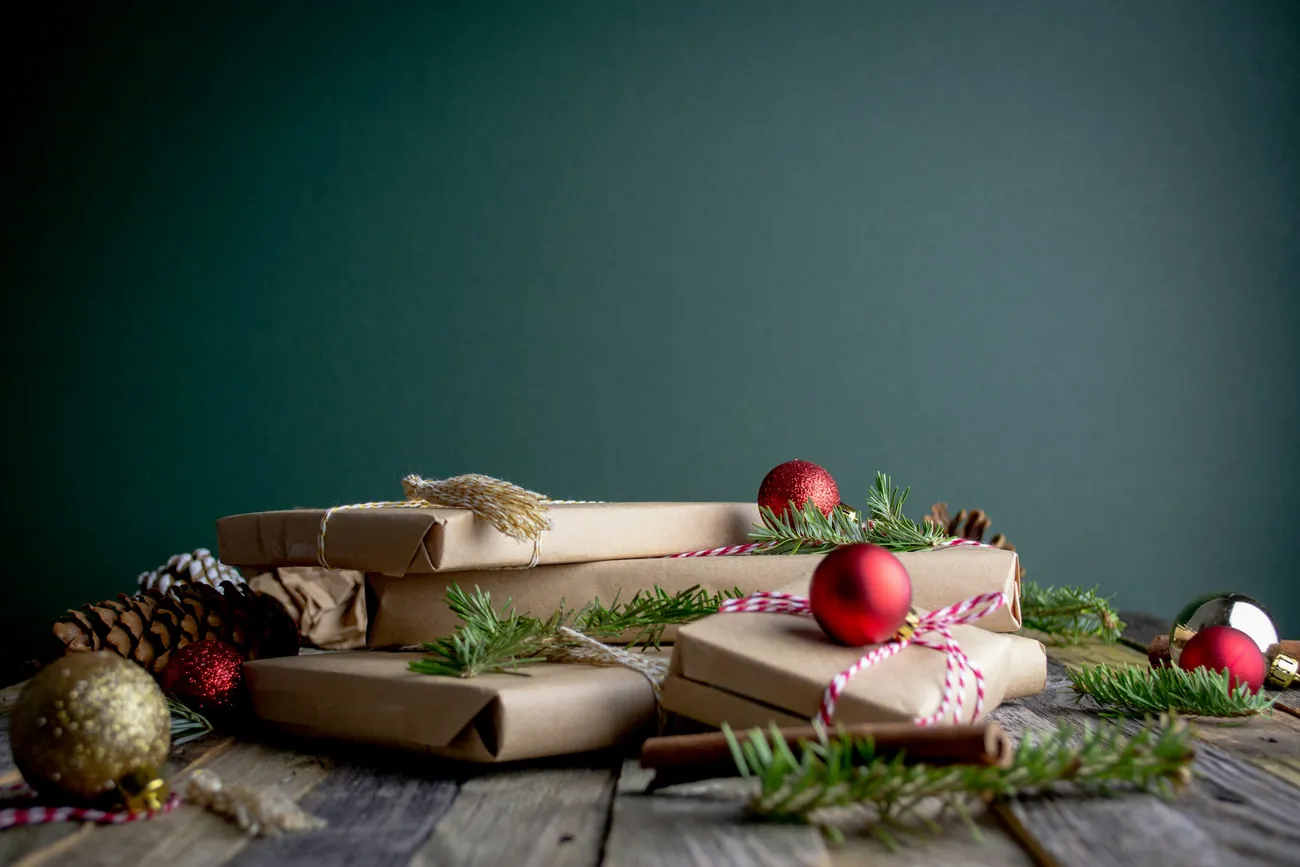 Gifts wrapped in brown paper and red-and-white string, with red baubles, pine cones and sprigs of fir on a wooden table.