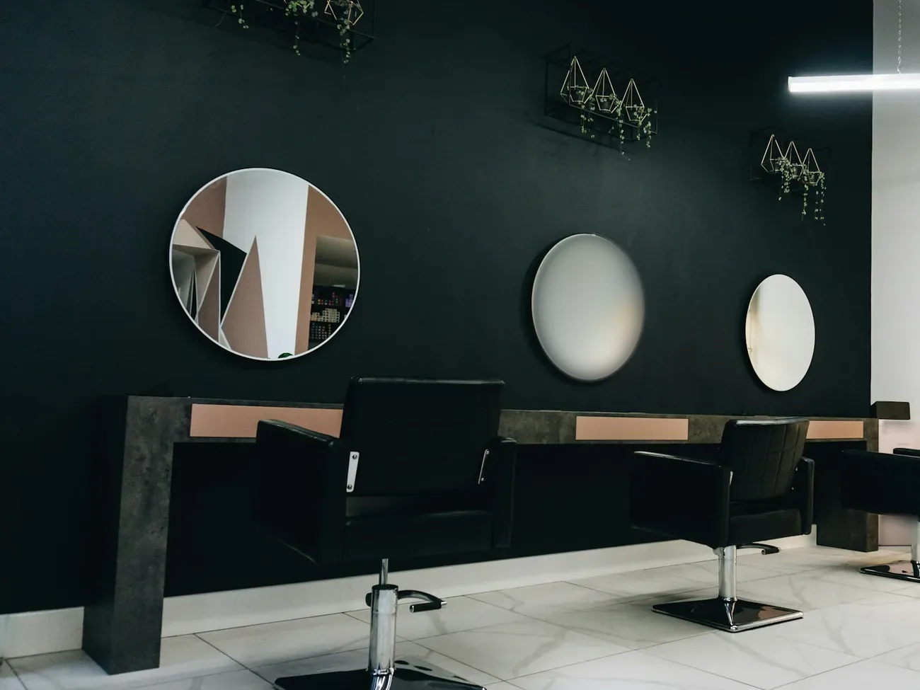 A salon with black styling chairs facing round mirrors on a dark wall, with small plants in brass holders above.