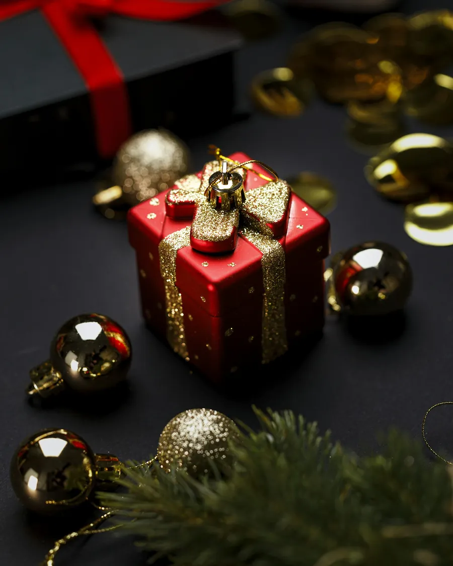 A small red gift box with gold ribbon, among baubles and pine.