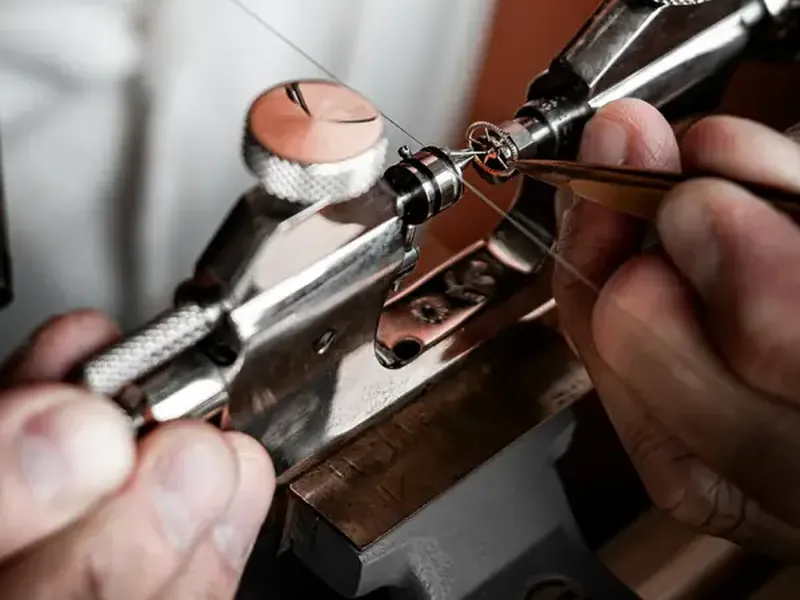 A watchmaker's hands at the bench, steadying a tiny part in a holder.
