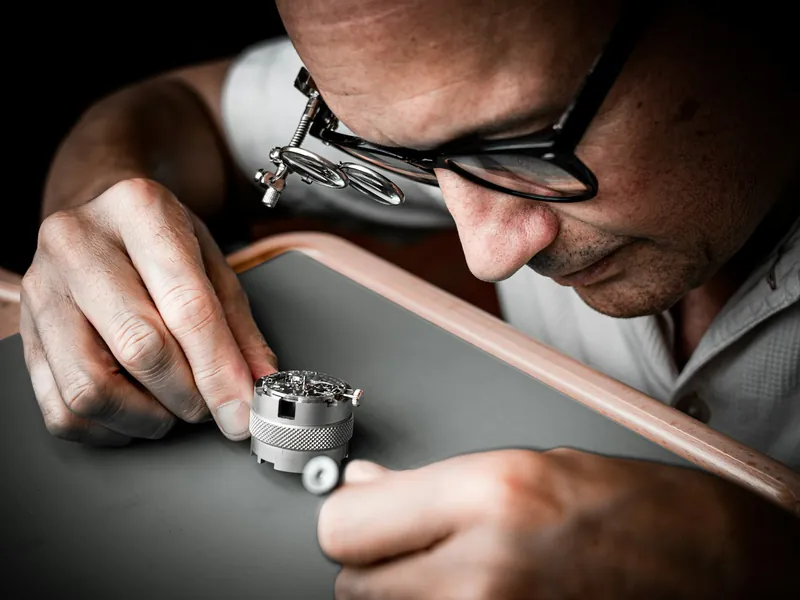 A watchmaker in glasses and a loupe peers closely at the workings of a watch.
